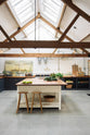 A large, renovated cattle shed has been transformed into a spacious pantry-blue Shaker kitchen, featuring an L-shaped cooker and sink run, along with a large white linen kitchen island topped with a stave hardwood worktop. Beautifully decorated with aged brass taps, Helen stools, luscious green vegetables, vintaged artisan ceramics, lilac tulips and abstract paintings.