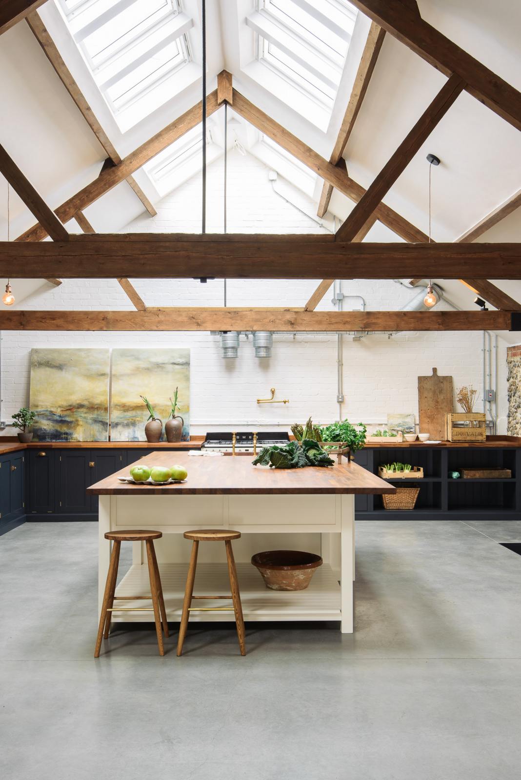A large, renovated cattle shed has been transformed into a spacious pantry-blue Shaker kitchen, featuring an L-shaped cooker and sink run, along with a large white linen kitchen island topped with a stave hardwood worktop. Beautifully decorated with aged brass taps, Helen stools, luscious green vegetables, vintaged artisan ceramics, lilac tulips and abstract paintings.