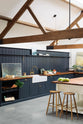 An oblique view of a large, renovated cattle shed has been transformed into a spacious pantry-blue Shaker kitchen, featuring an L-shaped cooker and sink run, along with a large white linen kitchen island topped with a stave hardwood worktop. Beautifully decorated with an aged brass Ionian tap, Helen stools, luscious green vegetables, vintaged artisan ceramics, lilac tulips and abstract paintings.
