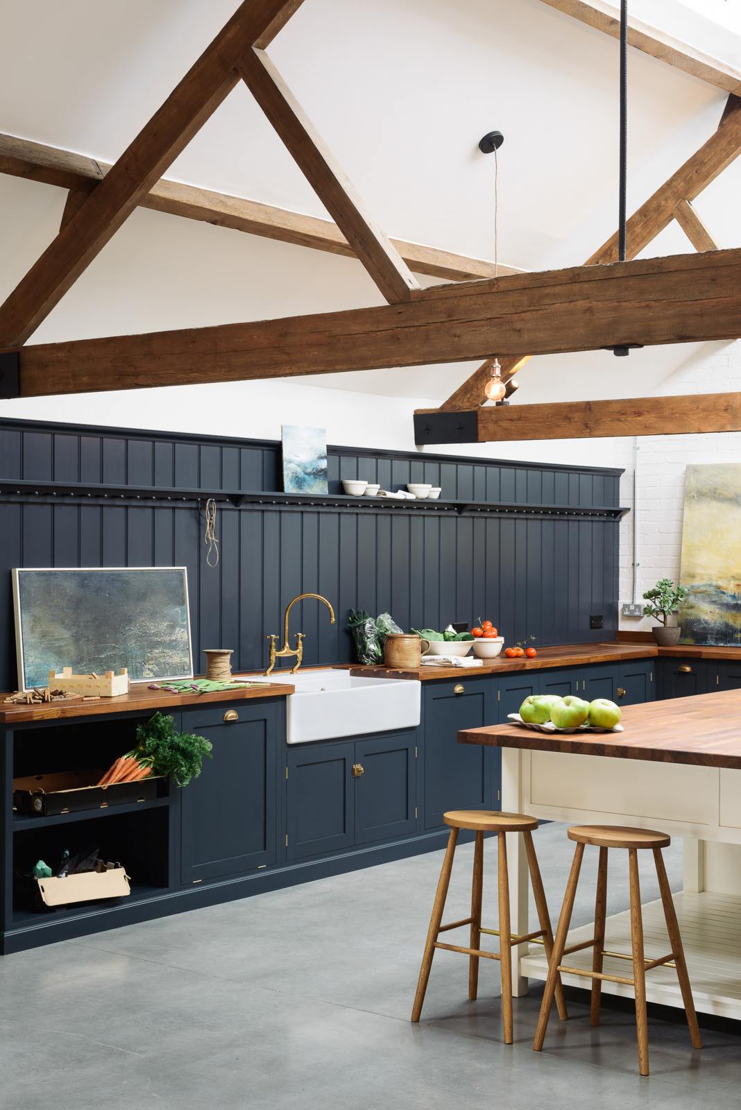 An oblique view of a large, renovated cattle shed has been transformed into a spacious pantry-blue Shaker kitchen, featuring an L-shaped cooker and sink run, along with a large white linen kitchen island topped with a stave hardwood worktop. Beautifully decorated with an aged brass Ionian tap, Helen stools, luscious green vegetables, vintaged artisan ceramics, lilac tulips and abstract paintings.