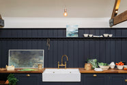 A large, renovated cattle shed has been transformed into a spacious pantry-blue Shaker kitchen, featuring an L-shaped cooker and sink run with a double Belfast sink, a long shelf with pegs, and a tongue-and-groove panel. Beautifully decorated with an aged brass Ionian tap, luscious green vegetables, vintage artisan ceramics, lilac tulips and abstract paintings.