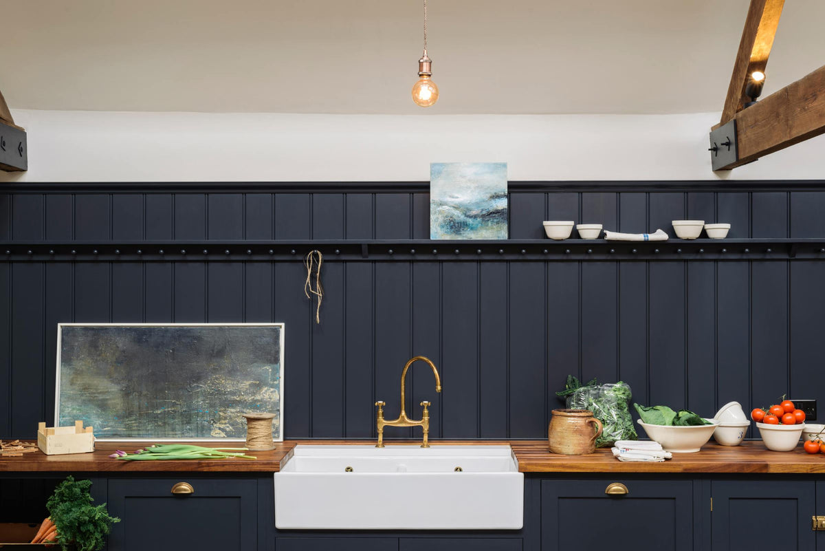 A large, renovated cattle shed has been transformed into a spacious pantry-blue Shaker kitchen, featuring an L-shaped cooker and sink run with a double Belfast sink, a long shelf with pegs, and a tongue-and-groove panel. Beautifully decorated with an aged brass Ionian tap, luscious green vegetables, vintage artisan ceramics, lilac tulips and abstract paintings.