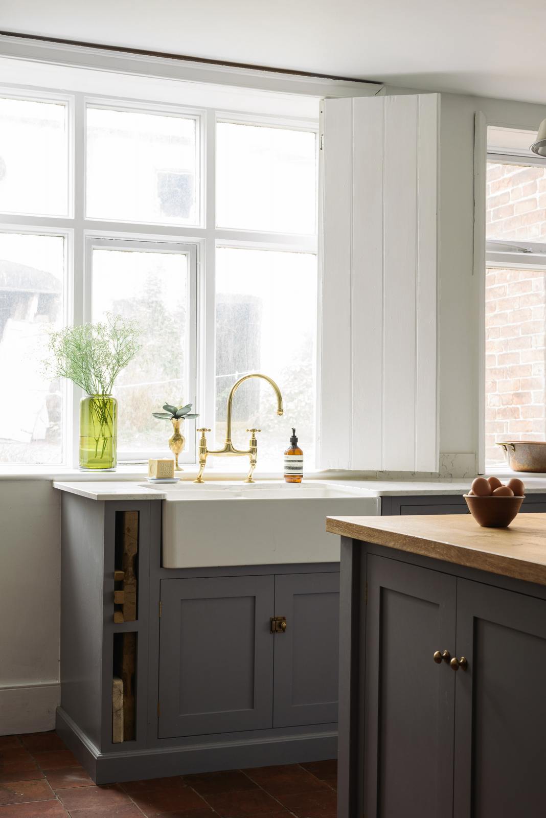 A closer look at a simple, modern shaker kitchen in a historic Cheshire townhouse, featuring a Silestone sink run with a Belfast sink and aged brass Ionian tap, and a large island with a solid oak worktop and a breakfast nook. Painted in muted cool grey with white walls and warm terracotta tile flooring. 
