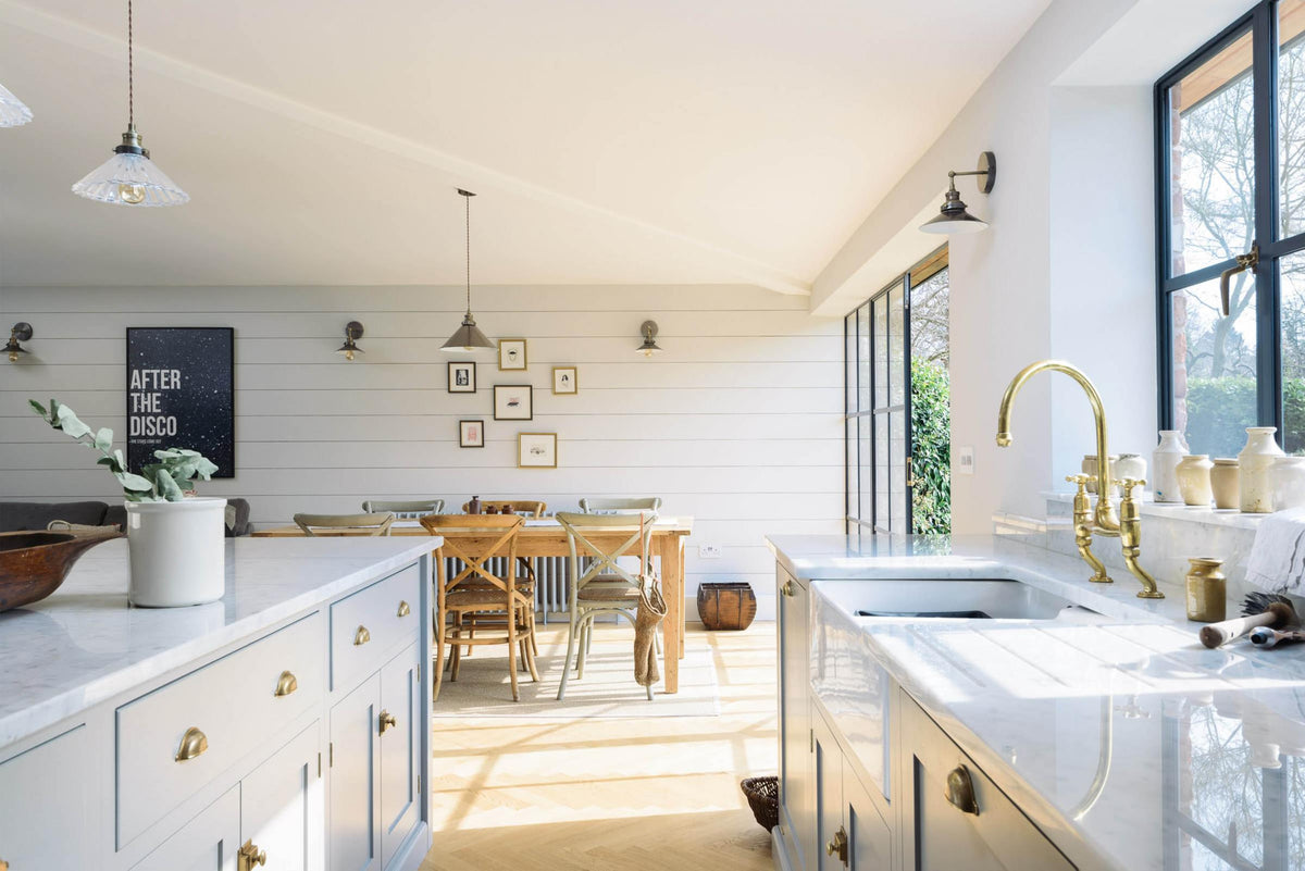 Nestled between a spacious and contemporary stone grey Shaker kitchen’s L-shaped sink and cooker run with a Carrara worktop, a single Belfast sink with an aged brass Ionian tap, and a kitchen island is a grand dining room with a long, solid oak dining table and chairs.