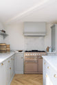 A corner of a spacious and contemporary stone grey Shaker kitchen features an L-shaped sink and cooker run with a Carrara worktop and splashback, a canopy, and a kitchen island. Elegantly decorated with artisan ceramics, a bowl of fresh eggs, and vintage baskets creates a cool and inviting space.