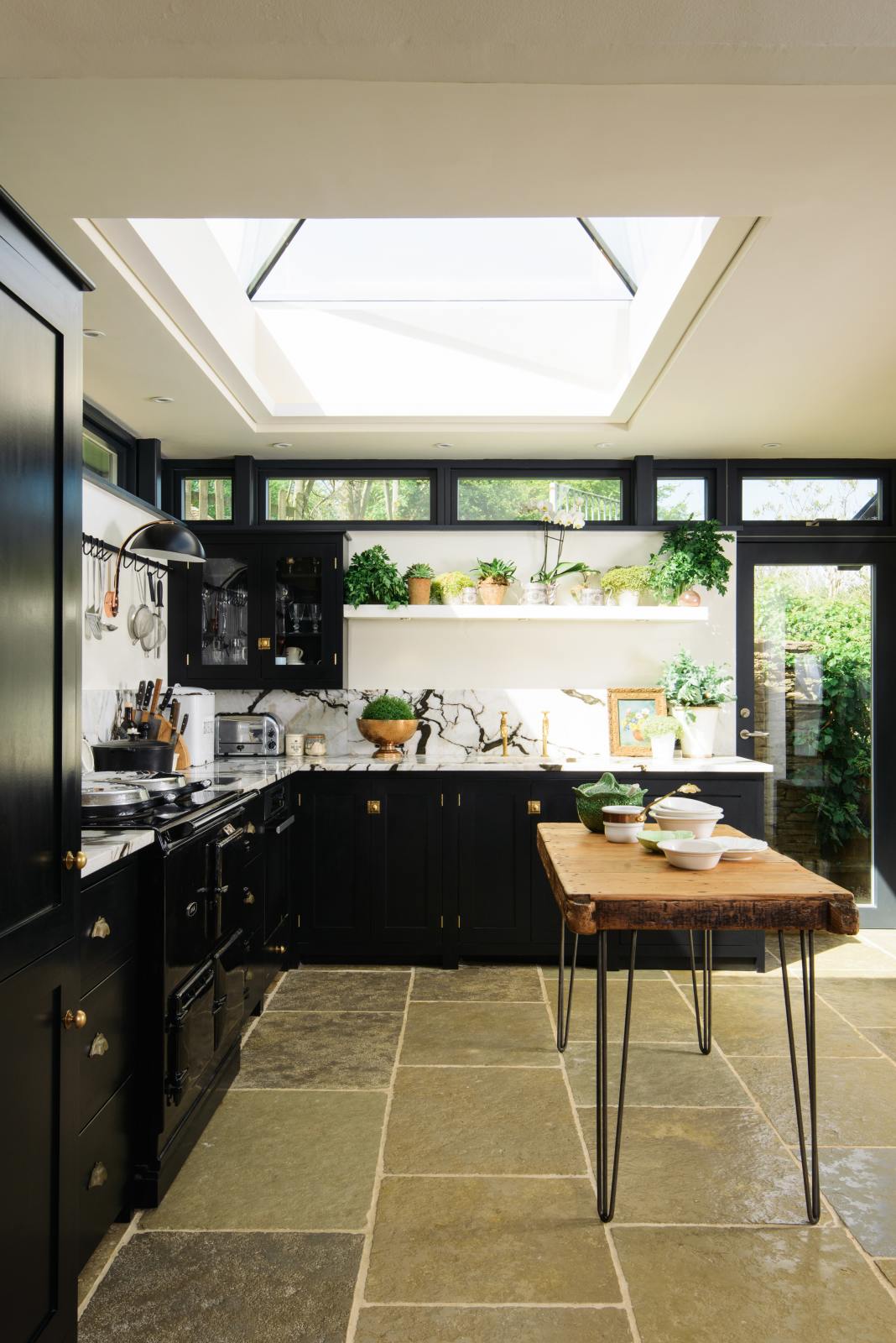 A straight view of a Painter’s Black shake kitchen with an L-shaped layout featuring a cooker and a sink. The worktop and splashback are crafted from San Simone quartzite. The design includes an overhead glazed cupboard and a tall storage cupboard, along with an aged brass hanging rail. The space is illuminated by a glass skylight, enhancing its overall brightness.
