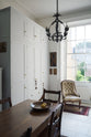 A close, oblique view of a tall yet simple Shaker kitchen featuring a sink and cooker run with a Carrara marble worktop, shelves, and a small bank of larder. Lovingly decorated with antique furniture, a vibrant red decorative rug, a variety of chic and curated paintings and artworks, and a vintage metal chandelier.