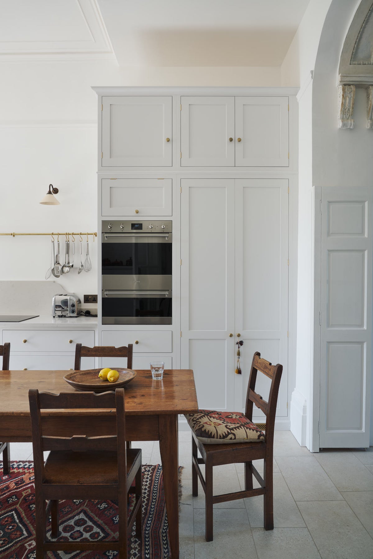 A practical corner of a tall yet simple Shaker kitchen featuring a sink and cooker run with a Carrara marble worktop, an oven cabinet, and an appliance cupboard. Lovingly decorated with antique furniture, a vibrant red decorative rug, a variety of chic and curated paintings, and a vintage metal chandelier.