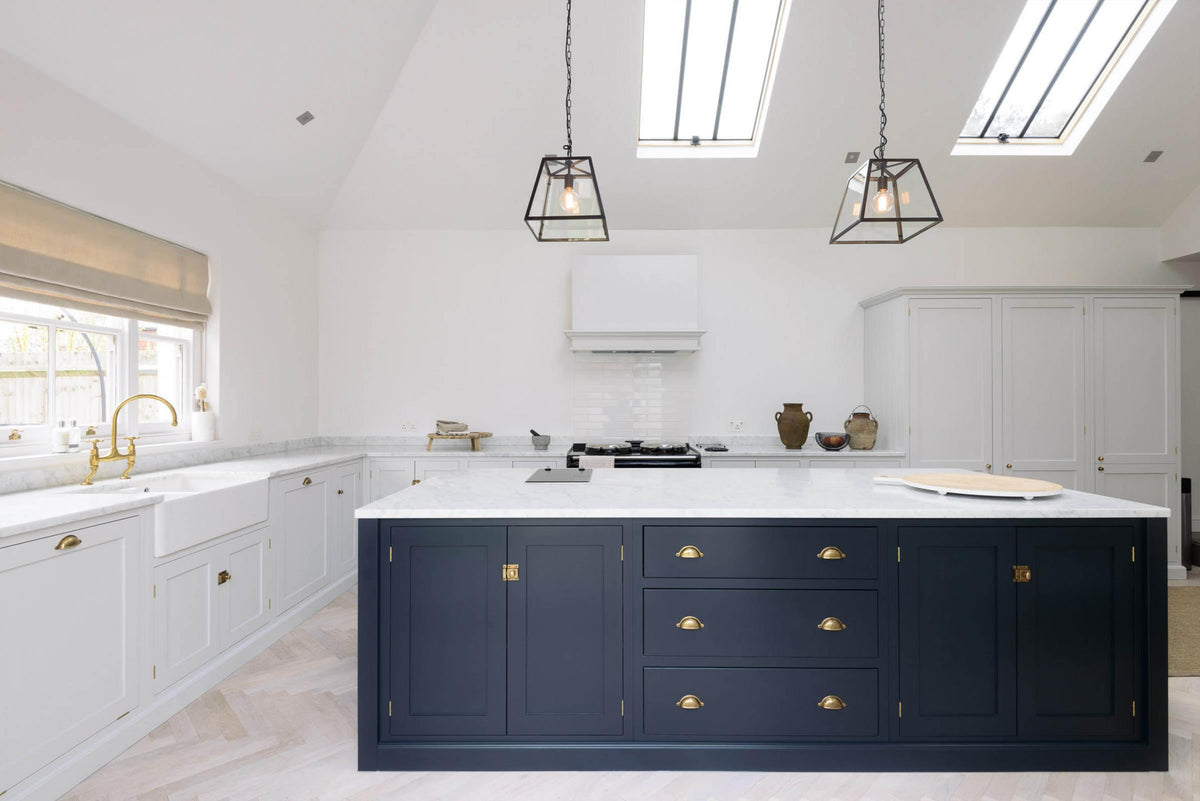 A spacious and artisan Shaker kitchen features an off-white L-shaped sink run with a double Belfast sink and an aged brass Ionian tap, a large pantry-blue kitchen island with a Carrara marble worktop, a canopy, white porcelain splashback tiles, and a bank of cupboards. Cleverly decorated with steel-framed hanging lights, vintage ceramics, and eclectic items serving as artistic displays, it creates a simple yet intriguing kitchen space.