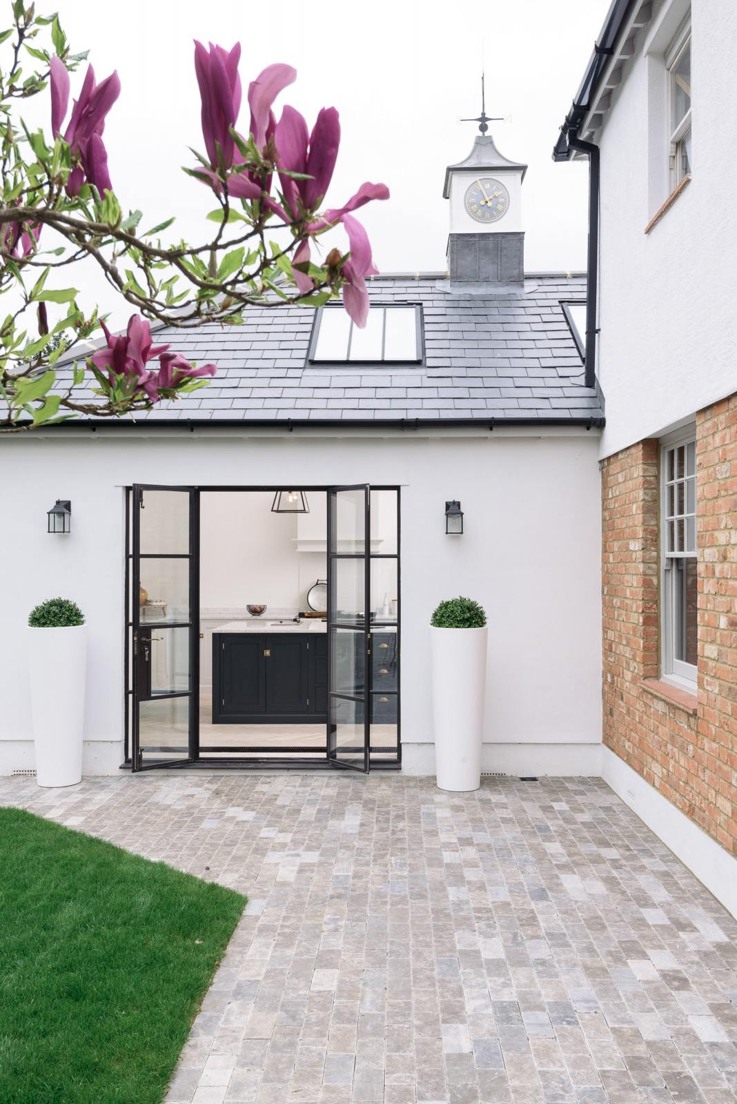 Outside the spacious and artisan Shaker kitchen is the modern and renovated exterior of the Victorian family home, featuring a pristine white wall mixed with the original brick texture, a black tiled roof, a small clock tower, a tiled pathway, freshly cut grass, and a bit of a fuchsia magnolia tree.