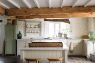 A simple English farmhouse kitchen in deVOL’s headquarters at Cotes Mill, with low, exposed wooden beams and worn wooden floorboards. The walls are textured and wobbly and painted in a light, off-white shade. The Shaker sink run, island and cooker run are all painted in Mushroom, a soft beige colour that feels timeless and classic.