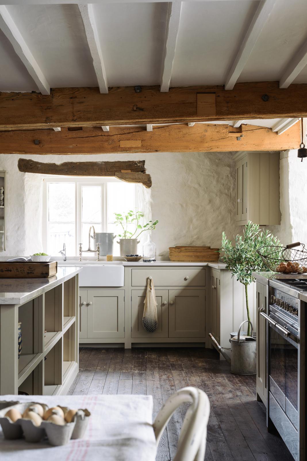 A sink run in a simple English farmhouse kitchen at deVOL’s headquarters, Cotes Mill. Low-exposed wooden beams and worn floorboards create a rustic atmosphere, while textured off-white walls add charm. The Shaker kitchen, including its island, is painted in Mushroom—a soft, timeless beige.