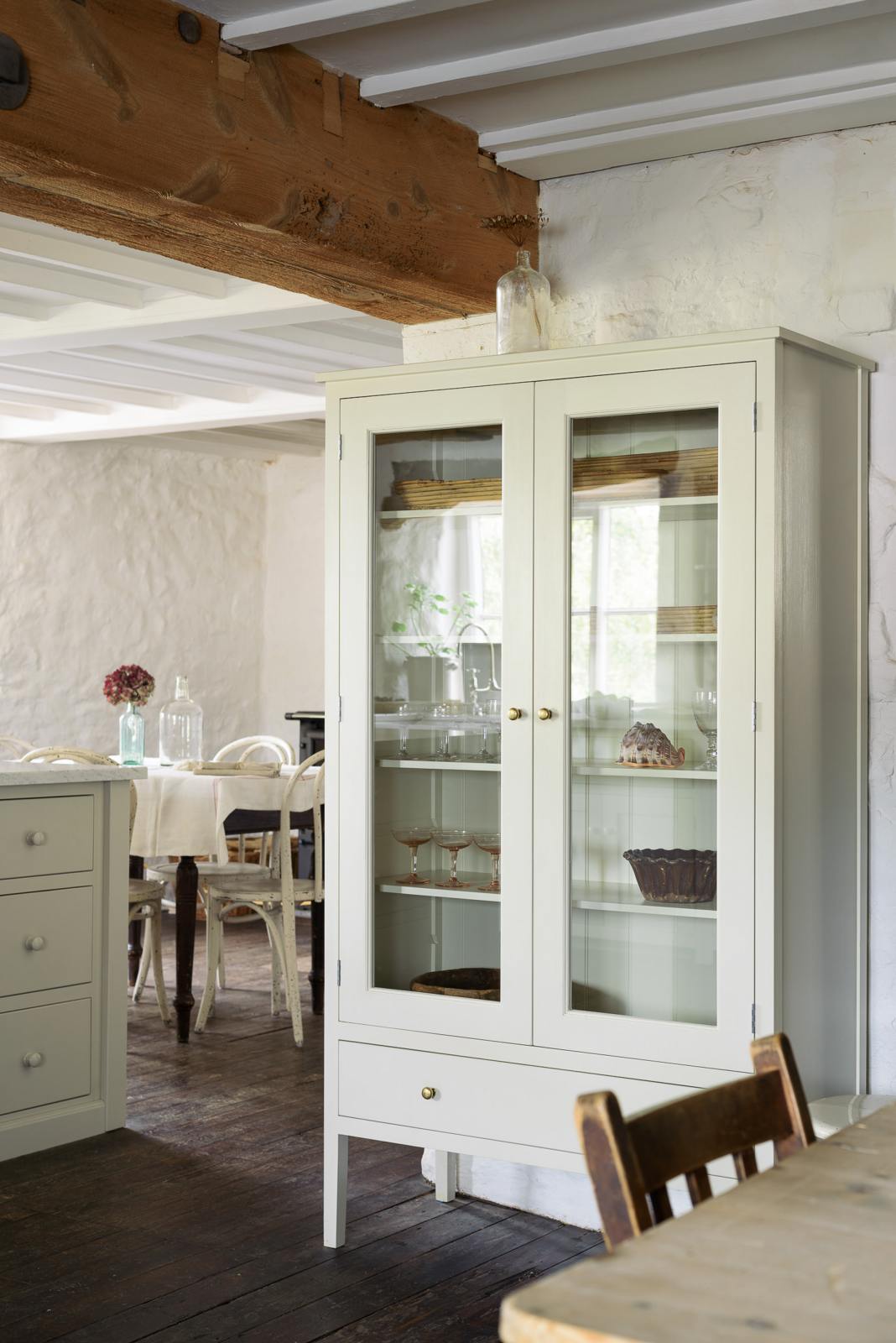 An angled shot of a small, free-standing cream cupboard, displaying assorted wine glasses, ceramics and a seashell. Part of the simple English farmhouse style kitchen in deVOL’s headquarters at Cotes Mill, with low, exposed wooden beams and worn wooden floorboards. Scattered is a collection of vintage items, including chairs, tables, and bric-a-brac, beautifully coordinated to complement the Mushroom Shaker kitchen.