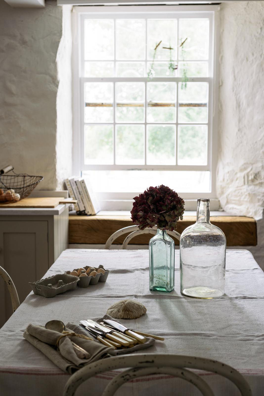 A moody scene with glasses, a carton of eggs, a seashell, and cutlery wrapped in napkins arranged on a white linen table, all bathed in soft natural light streaming through the window.