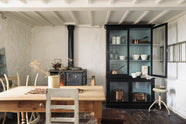 A simple English farmhouse kitchen in deVOL’s headquarters at Cotes Mill, with low, exposed wooden beams and worn wooden floorboards. The walls are textured and wobbly and painted in a light, off-white shade. There’s a deVOL custom cupboard, an iron-cast wood burner and vintage furniture.