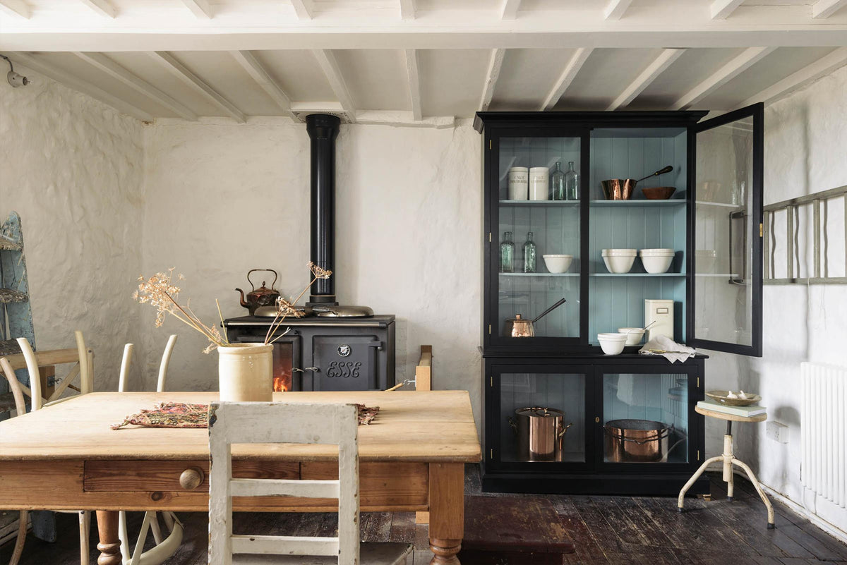 A simple English farmhouse kitchen in deVOL’s headquarters at Cotes Mill, with low, exposed wooden beams and worn wooden floorboards. The walls are textured and wobbly and painted in a light, off-white shade. There’s a deVOL custom cupboard, an iron-cast wood burner and vintage furniture.