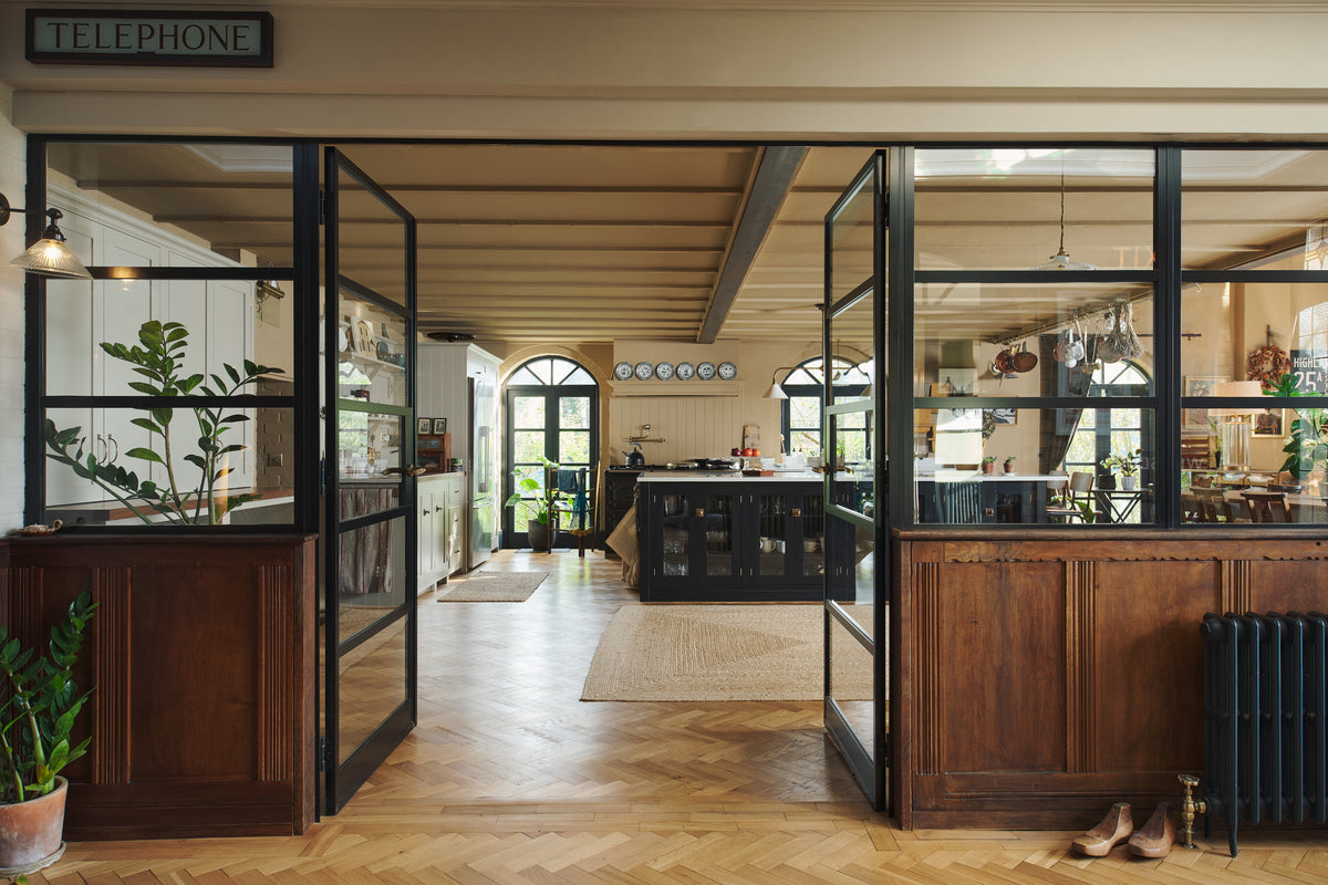 Past a pair of glass double doors lies a large, open Printer’s Black Shaker kitchen. A long cooker and sink run is topped with a Silestone worktop, complemented by both an open and closed pantry larder, and a kitchen island. Decorated with a show-stopping heirloom gaselier light and an aged brass pot filler, the room feels carefully considered, comfortable and highly functional.