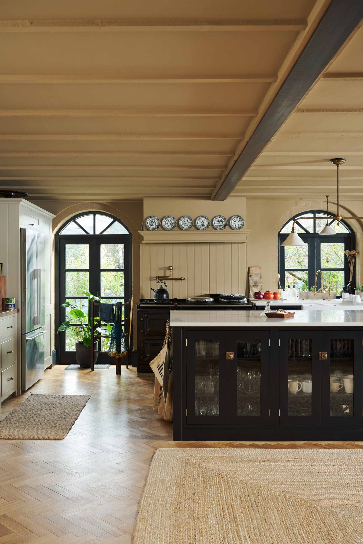 A large, open Printer’s Black Shaker kitchen. A long cooker and sink run is topped with a Silestone worktop, complemented by both an open and closed pantry larder, and a kitchen island. Decorated with a show-stopping heirloom gaselier light and an aged brass pot filler, the room feels carefully considered, comfortable and highly functional.