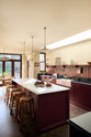 An oblique view of a simple yet elegant Refectory red shaker kitchen features a long L-shaped sink and cooker run with a black granite worktop, a kitchen island, a tongue and groove splashback, and a long shelf with brass rails. Beautifully decorated with artisan ceramics, antique champagne glasses, an oil painting, fresh produce, open skylights, and wooden parquet flooring, creating a bold yet understated kitchen space.