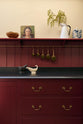 A close-up of the simple yet elegant Refectory red shaker kitchen features a long L-shaped sink and cooker run with a black granite worktop, a tongue and groove splashback, and a long shelf with brass rails. Beautifully decorated with artisan ceramics, antique champagne glasses, and an oil painting, creating a bold yet understated kitchen space.