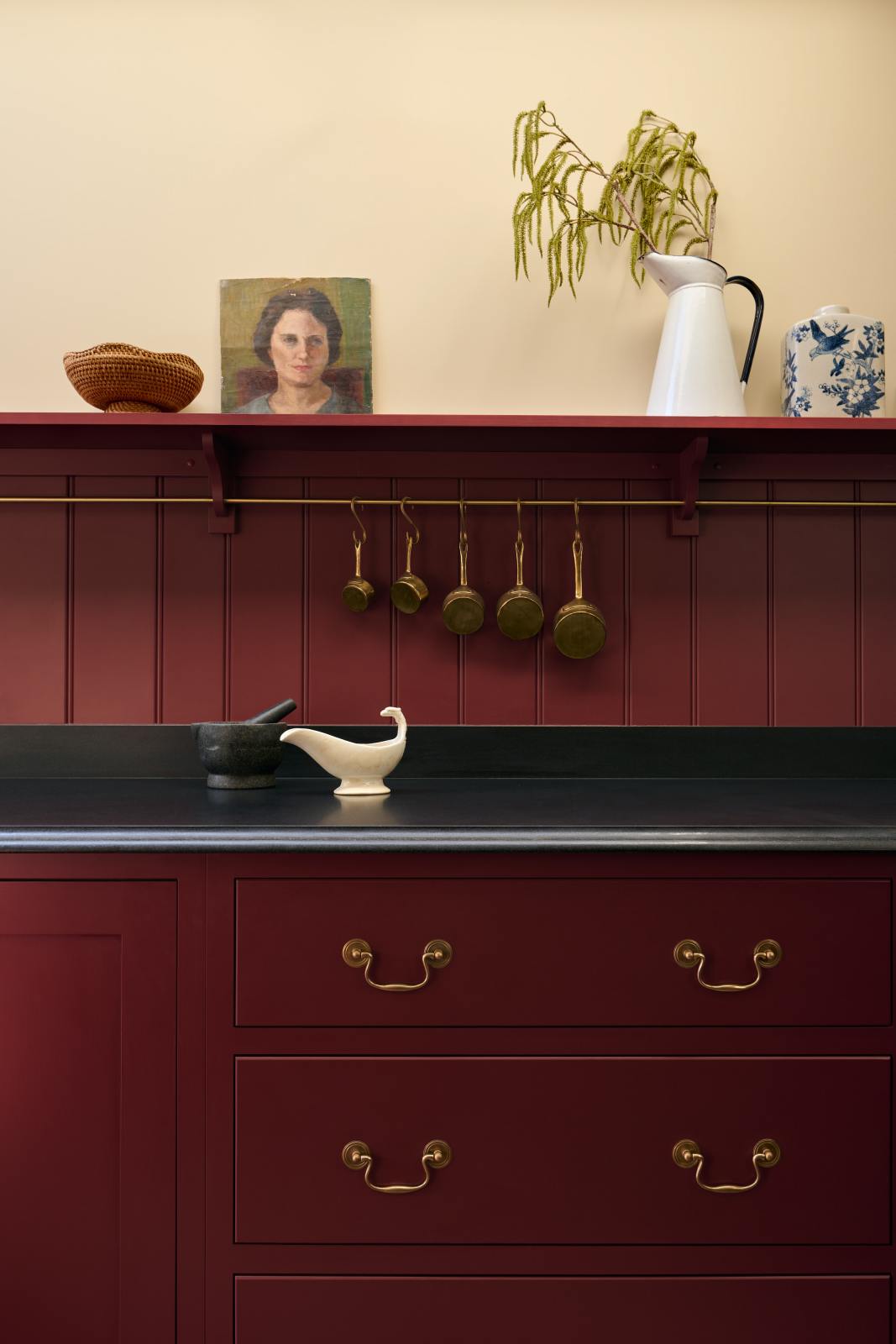A close-up of the simple yet elegant Refectory red shaker kitchen features a long L-shaped sink and cooker run with a black granite worktop, a tongue and groove splashback, and a long shelf with brass rails. Beautifully decorated with artisan ceramics, antique champagne glasses, and an oil painting, creating a bold yet understated kitchen space.
