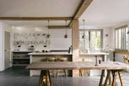 A tranquil and calm Shaker kitchen features a long sink and cooker run with a Silestone worktop and splashback, a single Belfast sink with a nickel Ionian tap, a long kitchen island with a breakfast nook, and shelves. Beautifully decorated with a vintage dining table that matches the raw wooden beams, muted ceramics, silver pendant lights, and a steel firewood basket, this kitchen creates a lovely, simple space that embraces natural materials.