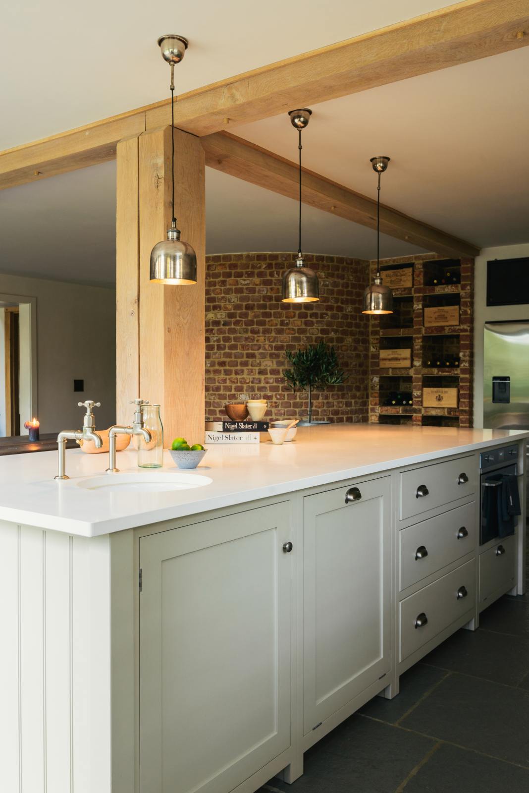 An oblique view of a tranquil and calm Shaker kitchen features a long kitchen island with a breakfast nook and a single sink with nickel Mayan taps. Beautifully decorated with silver pendant lights, a small bowl of limes, artisan ceramics, and a silver refrigerator, this kitchen creates a lovely, simple space that embraces natural materials.