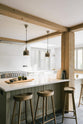 A quiet corner of a tranquil and calm Shaker kitchen features a long sink and cooker run with a Silestone worktop and splashback, a long kitchen island with a breakfast nook, and shelves. Beautifully decorated with vintage stools that match the raw wooden beams, muted ceramics, and silver pendant lights, this kitchen creates a lovely, simple space that embraces natural materials.