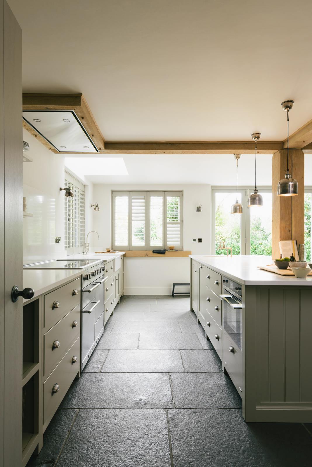 A tranquil and calm Shaker kitchen features a long sink and cooker run with a Silestone worktop and splashback, a single Belfast sink with a nickel Ionian tap, a long kitchen island with a breakfast nook, and shelves. Beautifully decorated with a chopping board that matches the wooden beams, some artisan ceramics, silver pendant lights, and an open book, this kitchen creates a lovely, simple space that embraces natural materials.