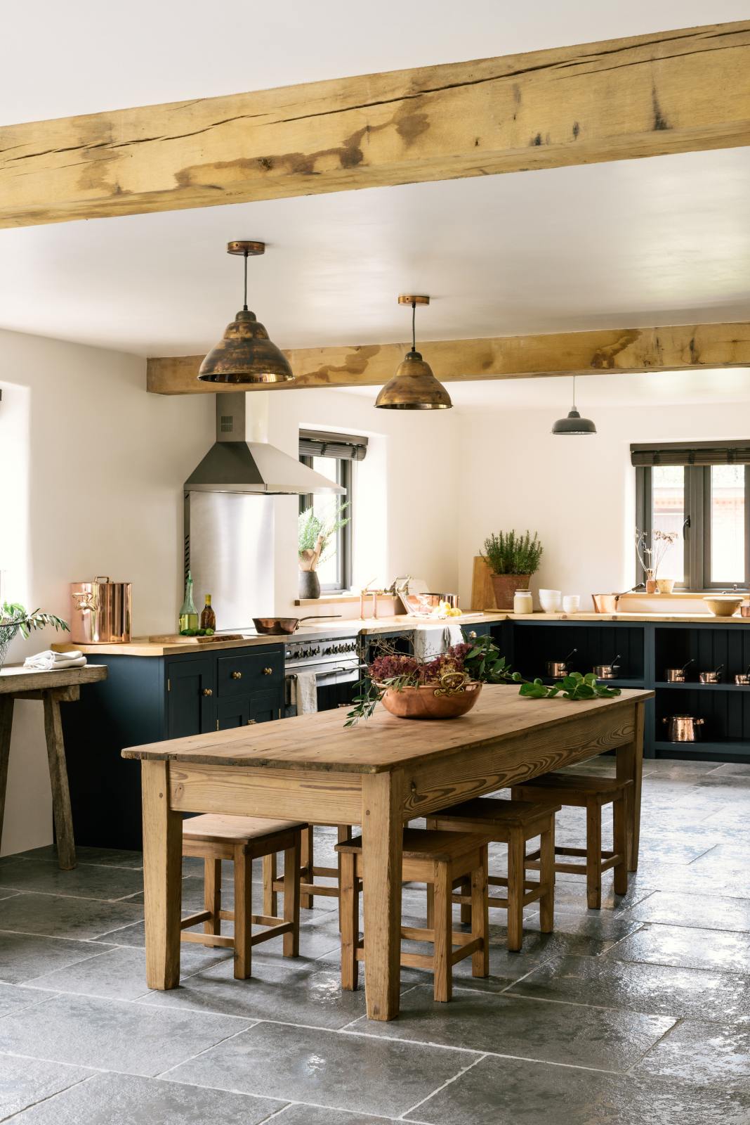 An angled view of a charming cottage shaker kitchen in Pantry Blue features a cooker run, open base cabinets, and a solid oak worktop that complements the wooden ceiling beams and the vintage dining table with stools.