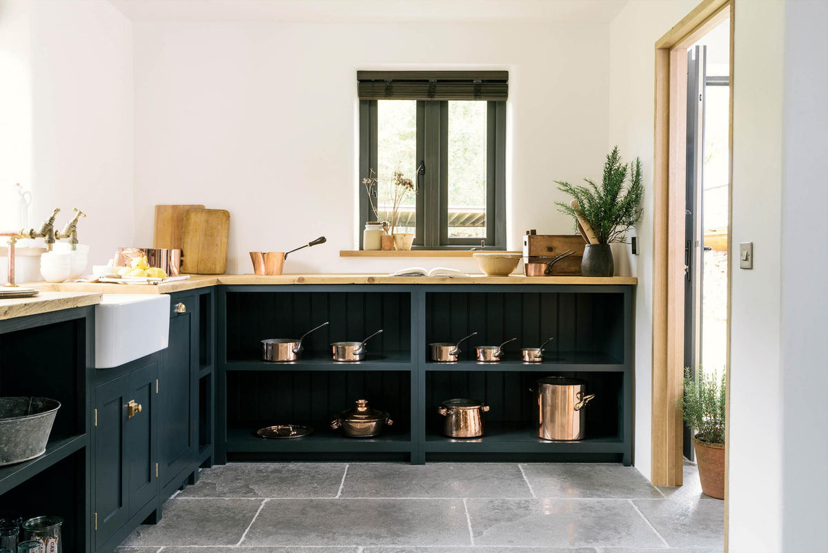 A closer look at the charming Pantry Blue cottage Shaker kitchen reveals a cooker run, open base cabinets, and a solid oak worktop that complements the wooden ceiling beams and vintage dining table with stools. The open cabinets showcase rose-gold cookware, reflecting the warmth of the wood while offering easy, functional access.