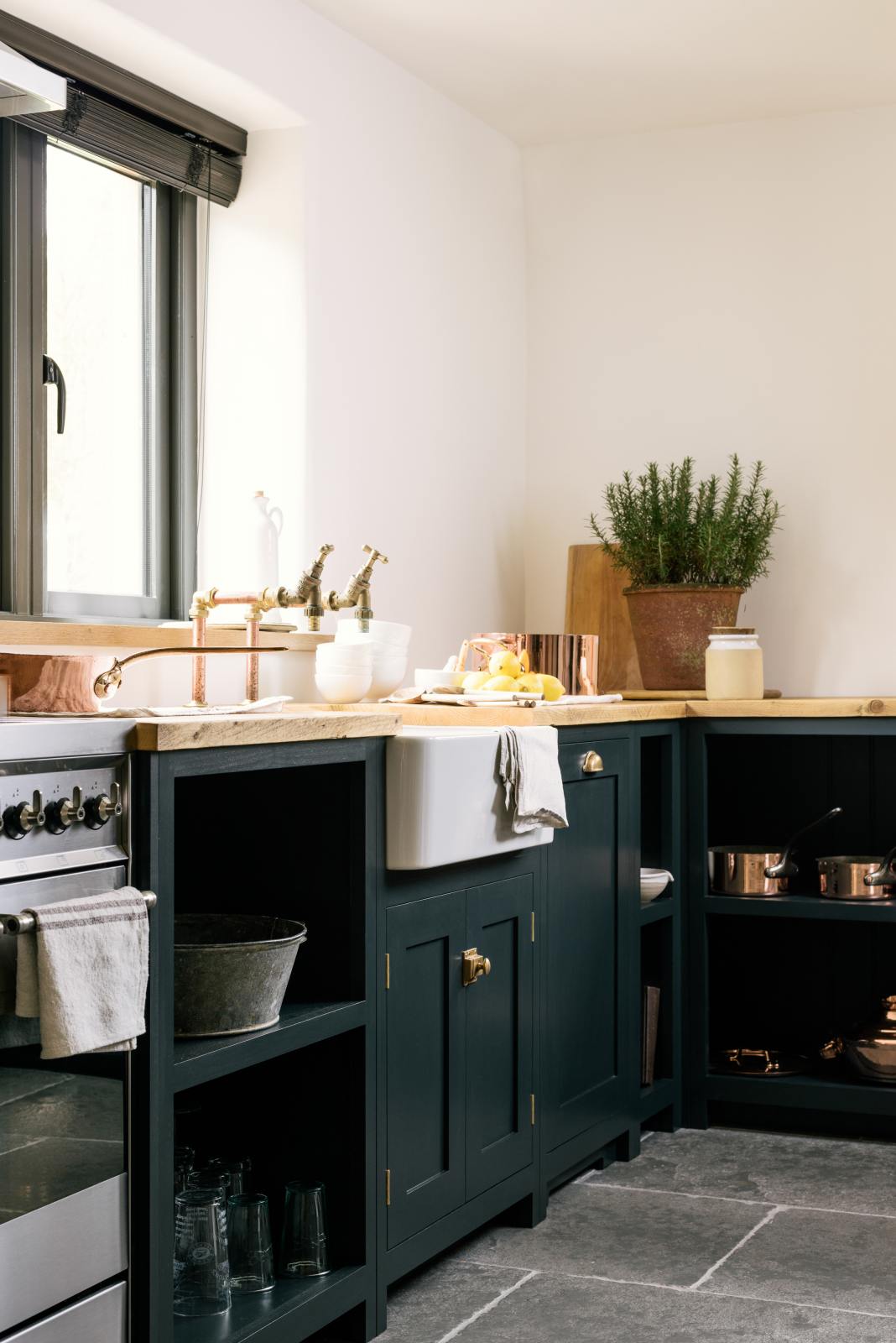 A closer look at the charming Pantry Blue cottage Shaker kitchen reveals a cooker run, open base cabinets, and a solid oak worktop that complements the wooden ceiling beams and vintage dining table with stools. The open cabinets showcase rose-gold cookware, reflecting the warmth of the wood while offering easy, functional access.