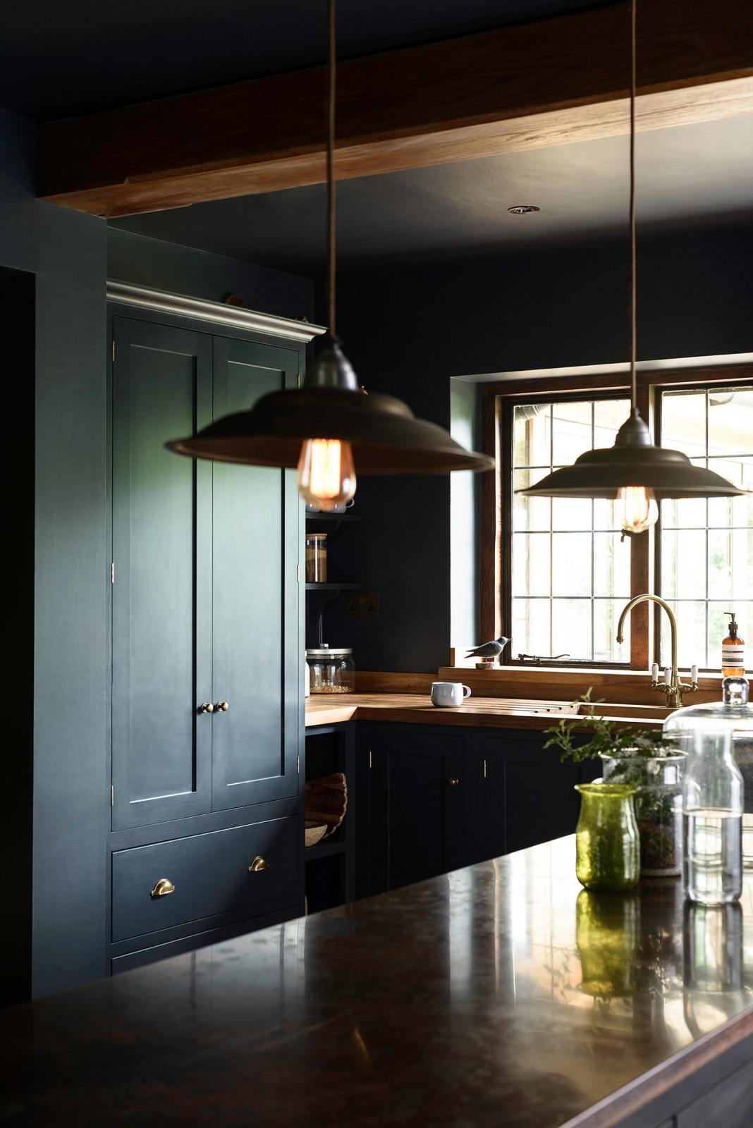 A dramatic view of a Pantry Blue shaker kitchen’s sink run and vintage island topped with Iroko wood. Light stone flooring enhances the space, featuring wooden ceiling accents, a large cooker splashback, and two hanging lights above the island.