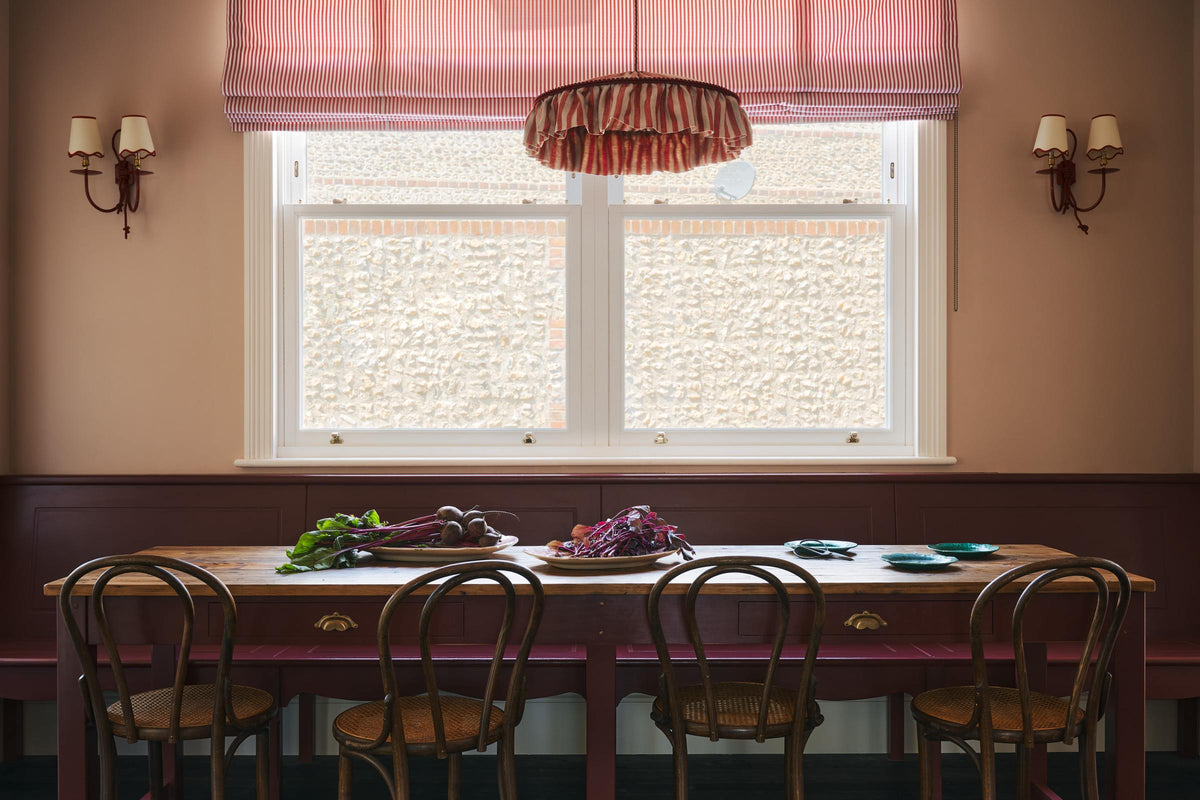 A grand, refectory red dining room with a Millhouse refectory table, Bentwood café chairs and a bench. The table is adorned with red beets and leaves, sea-green heirloom platters, a vintage red-and-white striped chandelier, and red-rimmed wall lights.