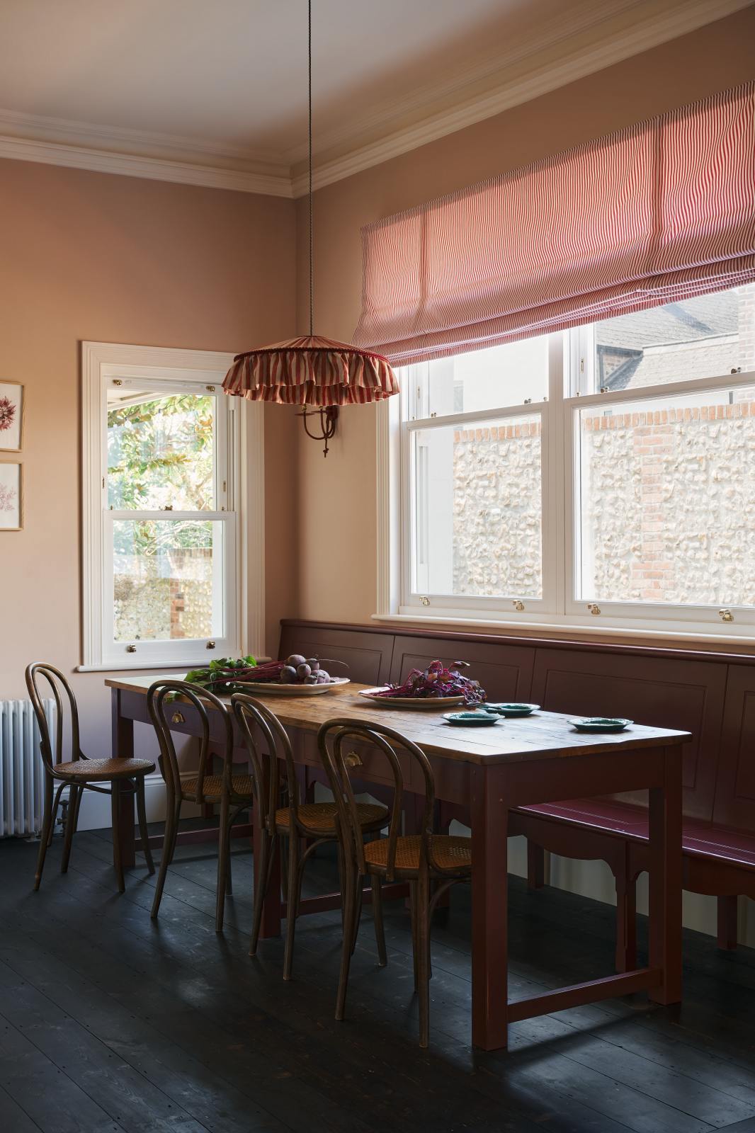 An angled view of a grand, refectory red dining room with a Millhouse refectory table, Bentwood café chairs and a bench. The table is adorned with red beets and leaves, sea-green heirloom platters, a vintage red-and-white striped chandelier, and red-rimmed wall lights.