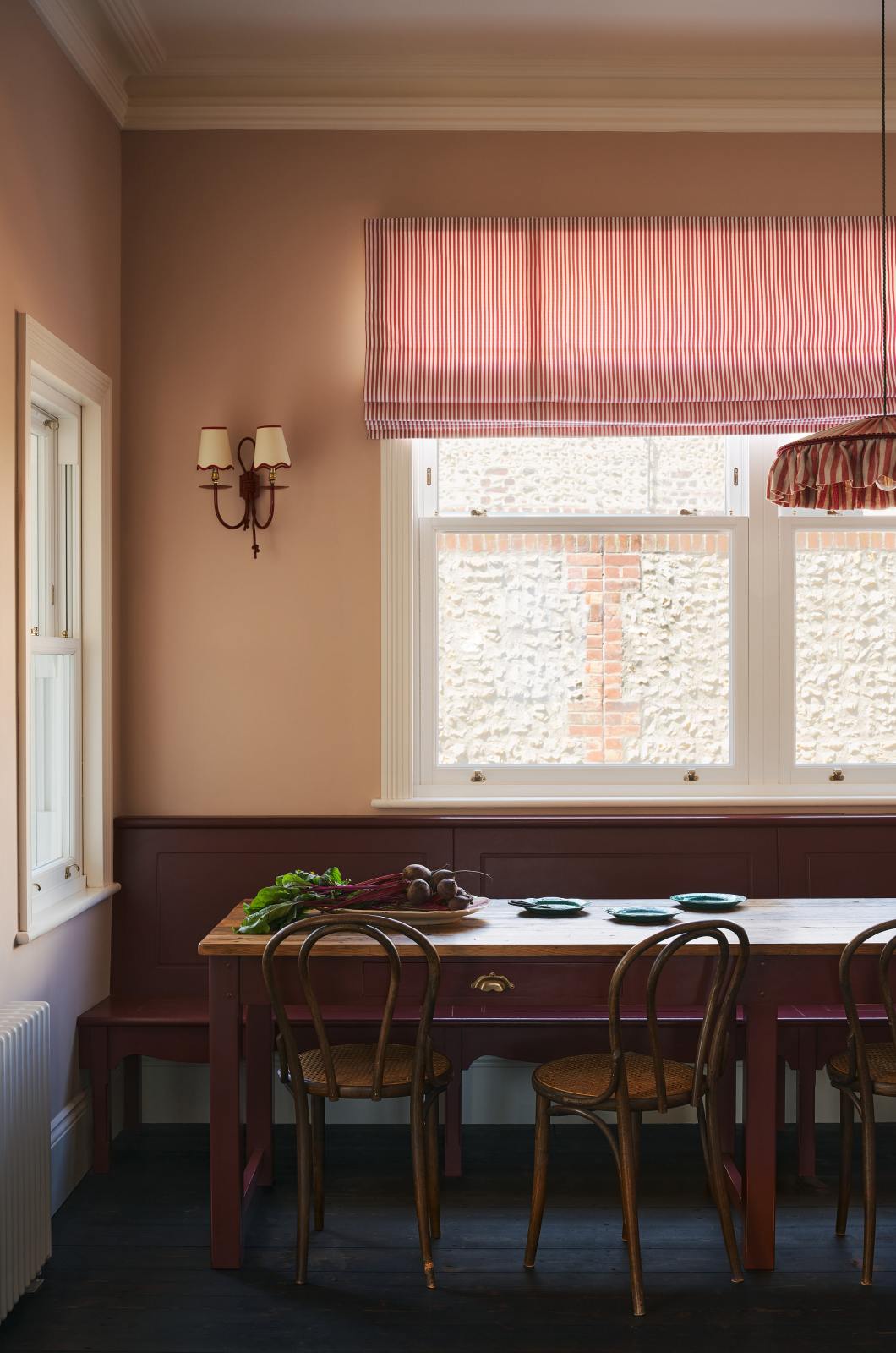 A corner of a grand, refectory red dining room with a Millhouse refectory table, Bentwood café chairs and a bench. The table is adorned with red beets and leaves, sea-green heirloom platters, a vintage red-and-white striped chandelier, and red-rimmed wall lights.
