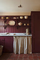 A humble refectory red laundry room features an L-shaped sink run with a Carrara marble worktop, shaker shelves with assorted vintage ceramics, towels, a full-height cupboard, and open cabinets covered by grid-patterned curtains on a rail.