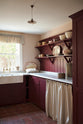 An angled view of a humble refectory red laundry room features an L-shaped sink run with a Carrara marble worktop, a double Belfast sink with an aged brass Ionian tap, open cabinets covered by grid-patterned curtains on a rail, shaker shelves with assorted vintage ceramics, and refreshing light from the window.