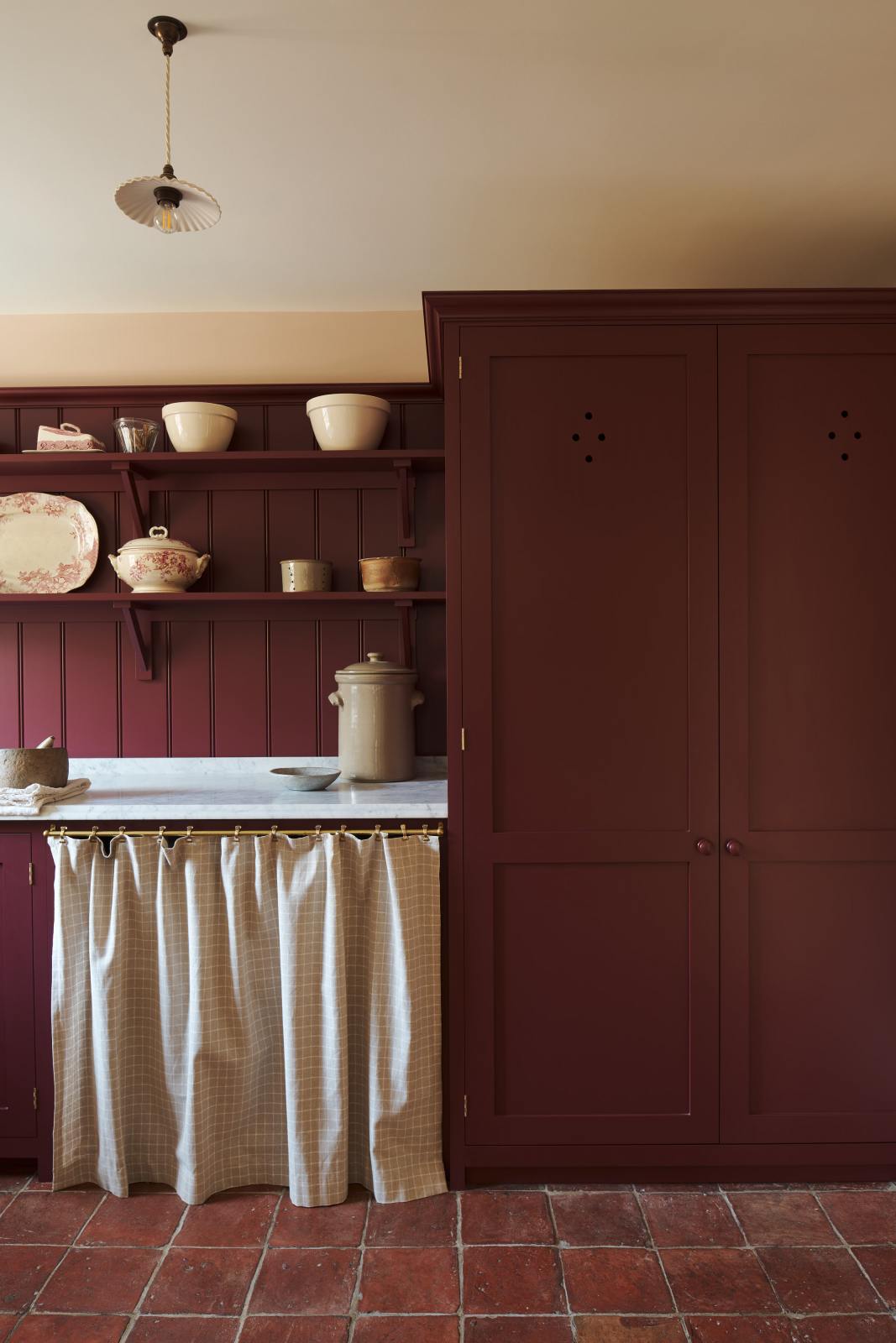 A humble refectory red laundry room features an L-shaped sink run with a Carrara marble worktop, shaker shelves with assorted vintage ceramics, towels, a large full-height cupboard, and open cabinets covered by grid-patterned curtains on a rail.