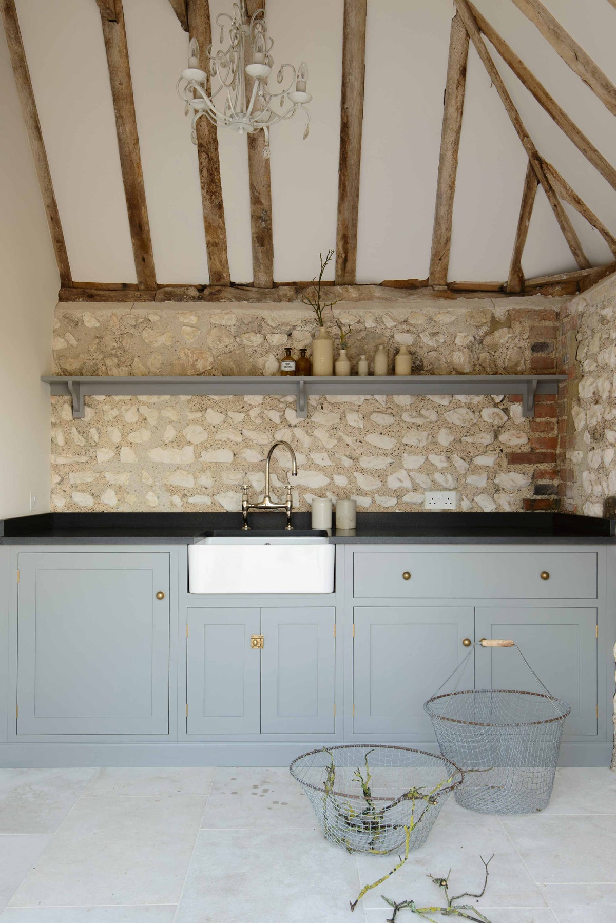 A chic and simple muted-blue sink run against the original wall textures of the pool house features a single Belfast sink with a nickel Ionian tap, a black honed granite worktop, and a long shelf. Adorned with vintage ceramics, brass fittings, and vintage steel baskets, it creates a functional corner in the space.