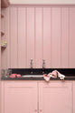 A closer look at a chic and simple muted-pink laundry room features a sink run with a honed black granite worktop, a full-height cupboard, a single sink, and nickel Mayan taps. Adorned with empty jam jars, folded tea towels, and a black washing machine, it creates a functional corner in the space.