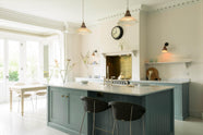 A minimalist Trinity Blue shaker kitchen includes a cooker around a chimney breast and an island with a Belfast sink in a bright, tall-ceilinged room. The kitchen is next to a small conservatory with its original French doors, allowing bright, natural light to fill the pale wooden breakfast table and cushioned chairs.