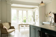 An angled view of the minimalist Trinity Blue shaker kitchen includes a cooker around a chimney breast and an island with a Belfast sink in a bright, tall-ceilinged room. The kitchen is next to a small conservatory with its original French doors, allowing bright, natural light to fill the pale wooden breakfast table, cushioned chairs and a French two-seater sofa.
