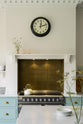 The central view of the minimal Trinity Blue shaker kitchen features a cooking area with a marble countertop, designed around a chimney breast, and complemented by a nearly golden-finish copper splashback. In the kitchen, you'll find a classic round clock with a black rim, assorted ceramics, glass vases filled with wildflowers, and a cream kettle sitting on the stove.