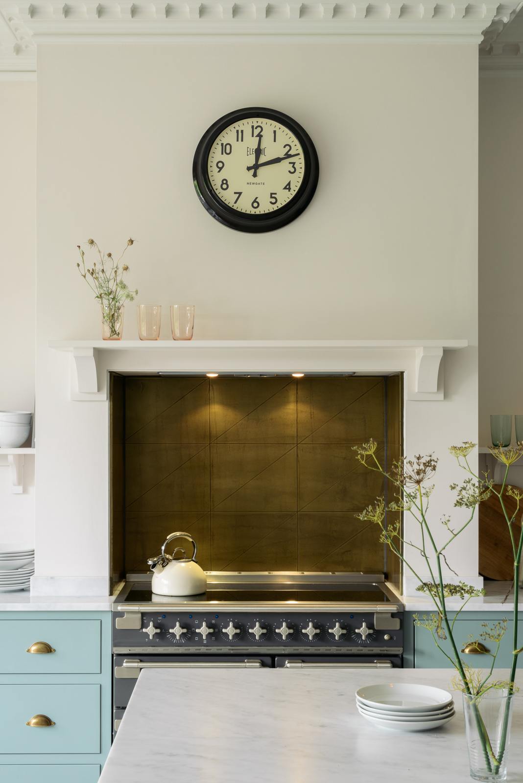 The central view of the minimal Trinity Blue shaker kitchen features a cooking area with a marble countertop, designed around a chimney breast, and complemented by a nearly golden-finish copper splashback. In the kitchen, you'll find a classic round clock with a black rim, assorted ceramics, glass vases filled with wildflowers, and a cream kettle sitting on the stove.