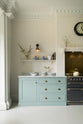 In a quiet corner of a minimal Trinity Blue shaker kitchen, the cooker run features a marble worktop. There are assorted ceramics, porcelain dishes, and books placed on the worktop and shelves.