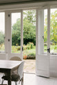 Across the pale wooden breakfast table and cushioned chairs, one of the French doors is open, inviting in the lovely view of green grass, trees, and a stone pathway. The bright colours of nature beautifully contrast with the light tones of the indoor space.