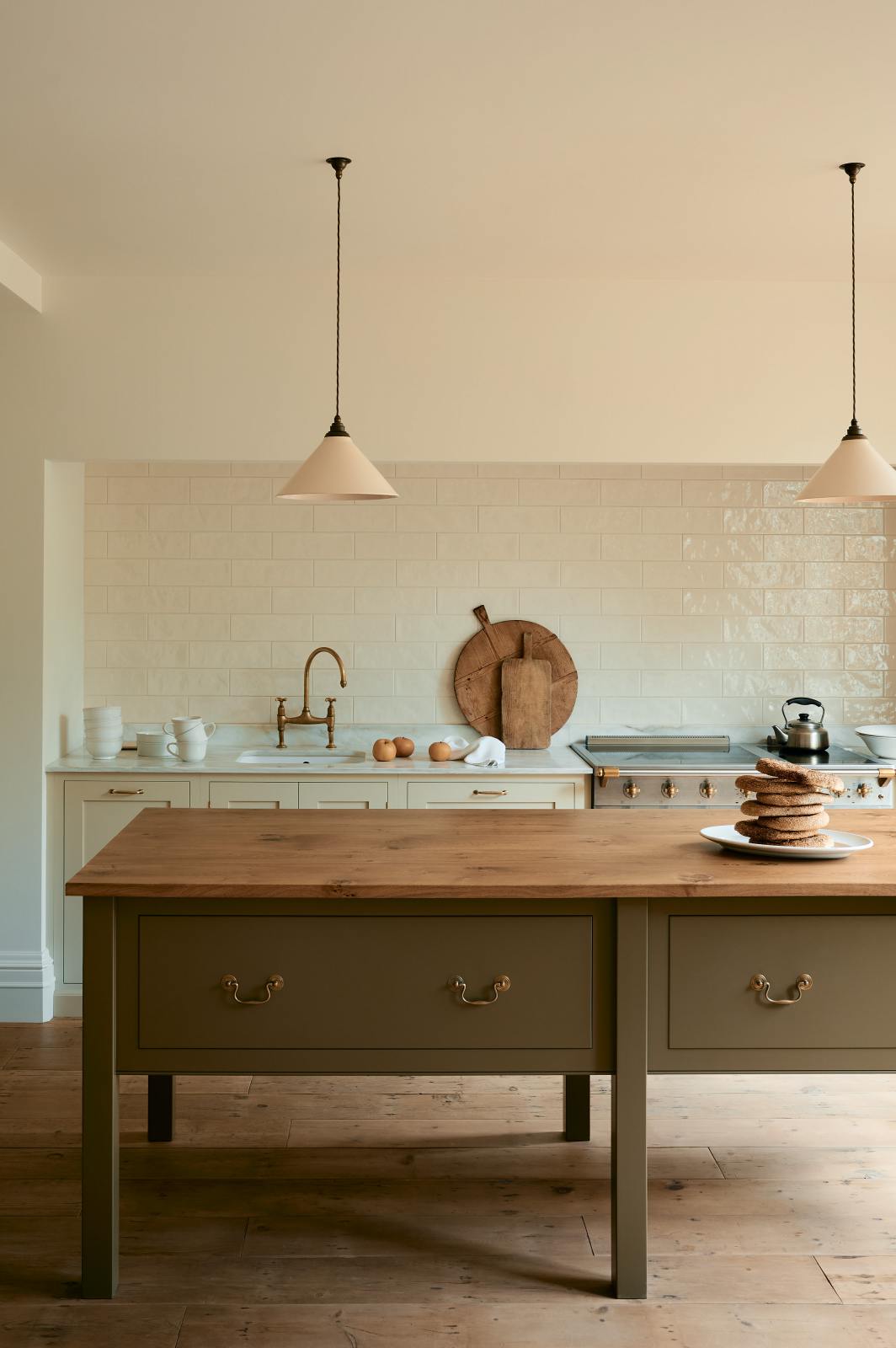 A closer look at a light, clean, and simple Shaker kitchen reveals a sink and cooker run nestled in a chimney breast, cream porcelain splashback tiles, wooden floorboards, and white walls. Cream cabinets complement an olive green Georgian-style prep table with deep drawers and a rustic oak worktop.