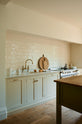 An angled and closer look at a light, clean, and simple Shaker kitchen reveals a sink and cooker run nestled in a chimney breast, cream porcelain splashback tiles, wooden floorboards, and white walls. Cream cabinets complement an olive green Georgian-style prep table with deep drawers and a rustic oak worktop.