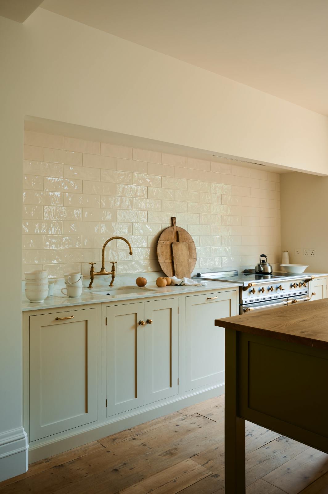 An angled and closer look at a light, clean, and simple Shaker kitchen reveals a sink and cooker run nestled in a chimney breast, cream porcelain splashback tiles, wooden floorboards, and white walls. Cream cabinets complement an olive green Georgian-style prep table with deep drawers and a rustic oak worktop.